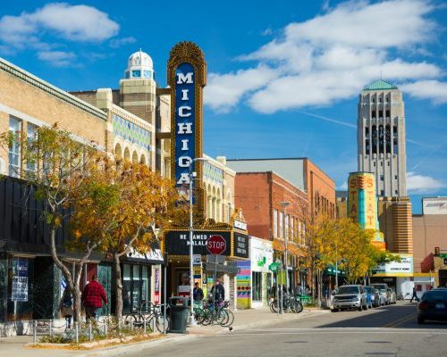 Ann Arbor, United States - October 18, 2015: People walking in the sidewalk of Liberty Street in Downtown Ann Arbor, with storefronts and the Michigan Theater sign, State Theater sign, cars and parked bicycles in the scene, and the Burton Memorial Tower in the distance, during a day with a blue sky with clouds.